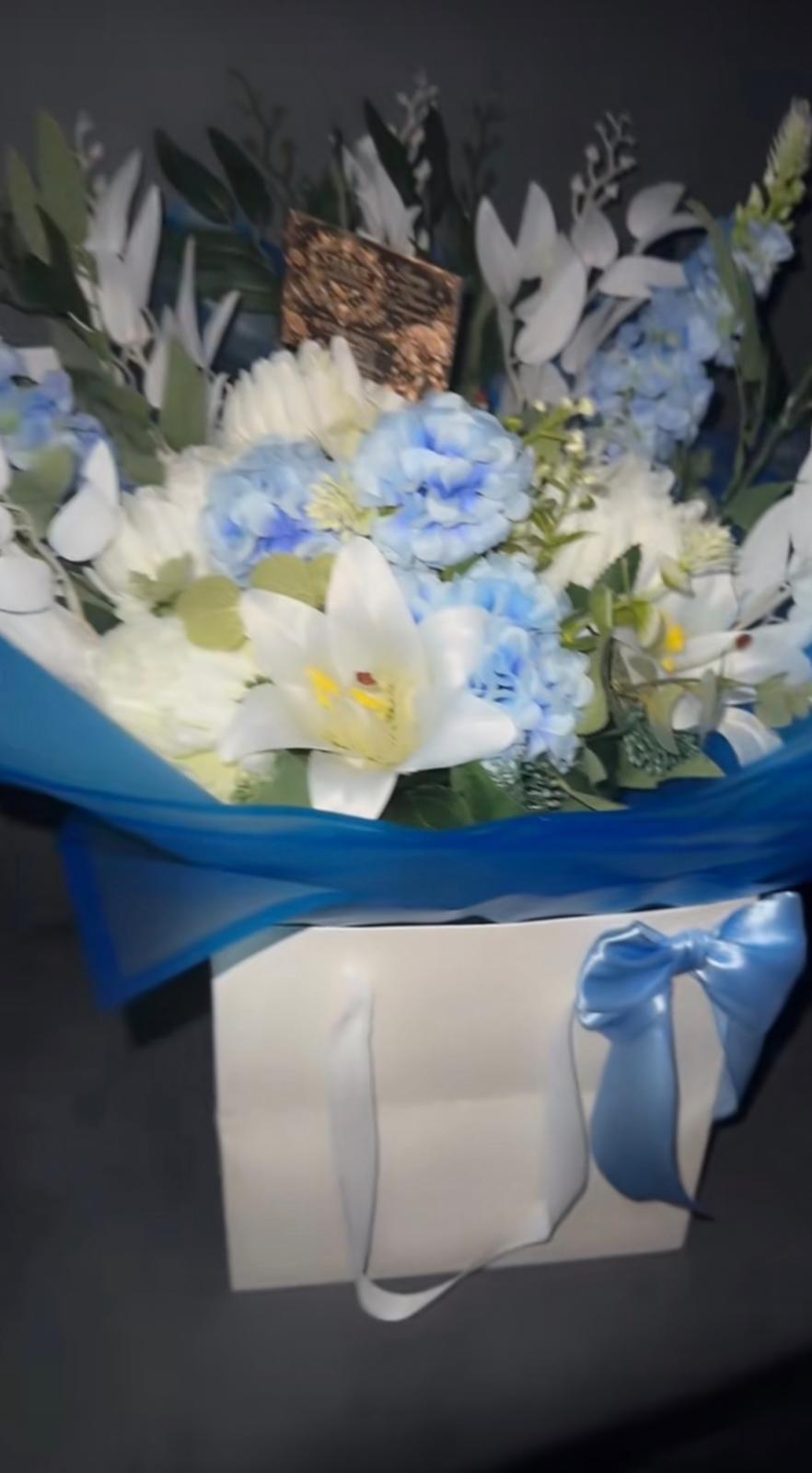White and blue artificial bouquet with lilies, hydrangea and delphinium in a white gift bag
